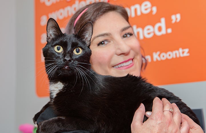 Smiling woman with a black cat with a white chest on her shoulder.