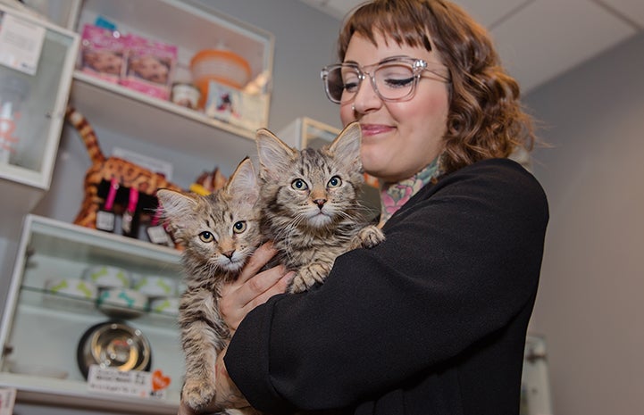 Smiling woman wearing glasses holding two brown tabby kittens in her arms